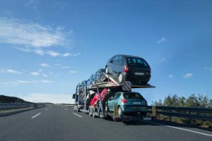 A car carrier truck from FL Auto Transport Clearwater transports several cars on a highway under a clear blue sky, with trees and guardrails alongside the road.