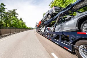 A car carrier truck, part of an FL Car Shipping Clearwater, FL fleet, transports multiple vehicles down a highway lined with trees and a concrete barrier on a clear day.