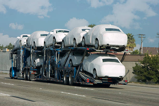 A car carrier truck transports several new cars covered in white protective wraps on a highway, exemplifying efficient Car Transport in Clearwater, FL, with trees and buildings in the background under a partly cloudy sky.