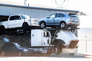 A car carrier truck is parked outside a building in FL, transporting three vehicles: a white pickup truck, a silver SUV, and a dark sedan. The vehicles are loaded on the truck’s trailer—ideal for Car Transport Clearwater needs—with one car partially inside the truck cab.
