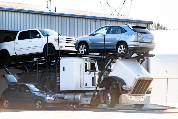 A car carrier truck is parked outside a building in FL, transporting three vehicles: a white pickup truck, a silver SUV, and a dark sedan. The vehicles are loaded on the truck’s trailer—ideal for Car Transport Clearwater needs—with one car partially inside the truck cab.