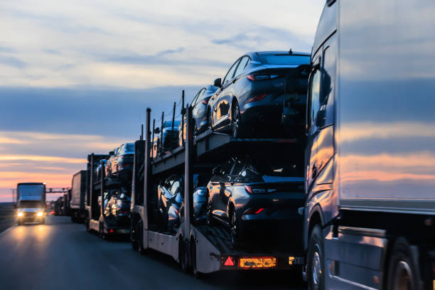 A car carrier truck loaded with new vehicles drives on a highway at sunset in FL, with other trucks visible in the background and headlights shining—showcasing the busy world of Car Transport Clearwater.